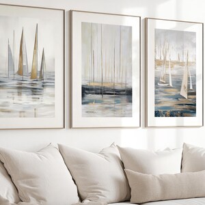 May include: Three framed abstract art prints showing sailboats on water. The artwork uses a muted palette of blues, greys, and gold. The frames are a light metallic colour, and the art is displayed above a cream-coloured sofa.