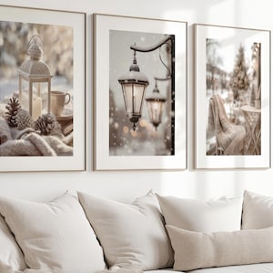 Op de afbeelding: Set van drie ingelijste prints met winterthema's. De prints tonen een lantaarn, met sneeuw bedekte straatlantaarns en een gezellige buitenscène met een deken en een stoel. Het kleurenpalet is neutraal met zachte wittinten, crèmes en bruinen.