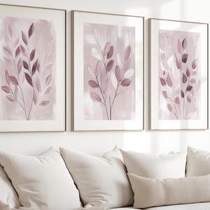 May include: A set of three framed wall art prints featuring abstract botanical illustrations of leafy branches in shades of dusty rose, mauve, and white. Each rectangular print is encased in a thin, light-colored frame, displaying a soft, painterly background with delicate, dark-lined foliage extending upward.