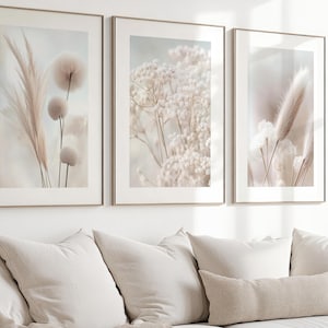Könnte beinhalten: Drei gerahmte botanische Drucke in neutralen Tönen. Das Kunstwerk zeigt getrocknete Blumen und Gräser in Beige- und Cremetönen. Die Rahmen haben eine goldene Oberfläche. Die Drucke werden über einem beigen Sofa mit weißen Kissen angezeigt.
