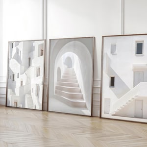 May include: Three framed wall art prints featuring white architectural scenes. The left print shows stacked white buildings, the centre print displays a white arched staircase, and the right print depicts a white building facade with stairs and doors.