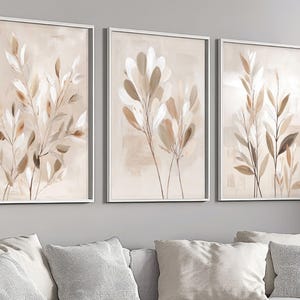 May include: A set of three vertical wall art prints in white frames, each featuring abstract botanical illustrations of leaves and stems in shades of beige, cream, and soft brown against a neutral, light-colored background.