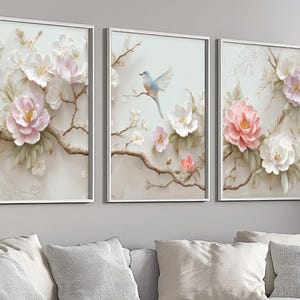 Pode incluir: Conjunto de três peças de arte de parede emolduradas com desenhos florais e de pássaros. Cada peça tem uma moldura branca e retrata galhos com flores brancas e rosas. Um painel mostra um pássaro azul.