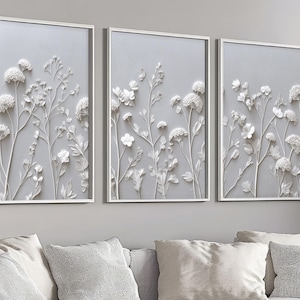 Può includere: Tre opere d'arte da parete incorniciate con disegni floreali bianchi su sfondo grigio. L'opera d'arte ha un effetto rilievo 3D, con dettagliate raffigurazioni di fiori e fogliame. Le cornici sono bianche e l'estetica generale è moderna ed elegante.