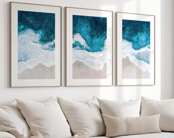Può includere: Un set di tre stampe artistiche da parete incorniciate verticalmente che mostrano una vista aerea di una spiaggia sabbiosa con onde turchesi che si infrangono sulla riva. Ogni stampa è racchiusa in una cornice sottile e chiara, con acqua blu.
