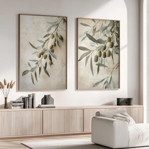 May include: Two framed prints of olive branches with green olives on a light beige background. The prints are hanging on a white wall above a light wood cabinet.