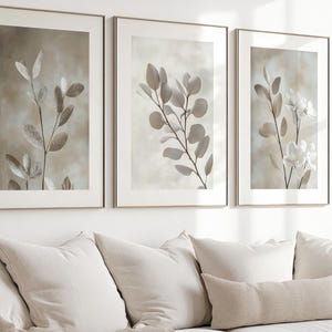 May include: A set of three framed wall art prints featuring botanical illustrations of eucalyptus branches and white flowers in soft beige and neutral tones, displayed in thin light-colored frames against a plain white wall.