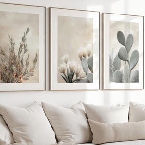 以下が含まれることがあります： 植物の画像が描かれた3枚の額入りウォールアートプリントのセット。左のプリントは乾燥した草、中央は花を咲かせたサボテン、右はウチワサボテンを描いており、すべて落ち着いたベージュとグリーンの色調で統一されています。