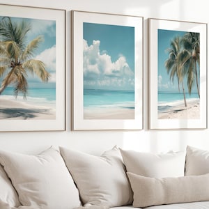 May include: Three framed art prints depicting a tropical beach scene. Each print features a palm tree, white sand, and turquoise water under a blue sky with clouds. The frames are a light metallic color, and the prints are displayed above a beige sofa.
