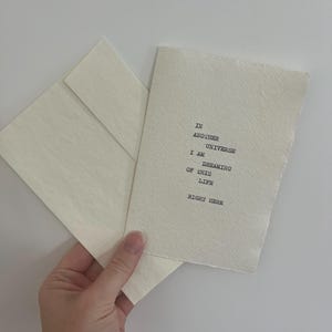 May include: A cream-coloured textured paper card with a deckled edge featuring black typewriter-style text that reads: IN ANOTHER UNIVERSE I AM DREAMING OF THIS LIFE RIGHT HERE. The card is accompanied by a matching cream-coloured envelope.