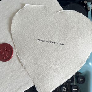 Könnte beinhalten: Eine weiße Keramik-Tasse mit schwarzem Henkel und dem Text "But First, Coffee" in schwarzer, fetter Schrift. Die Tasse steht auf einer weißen Oberfläche.