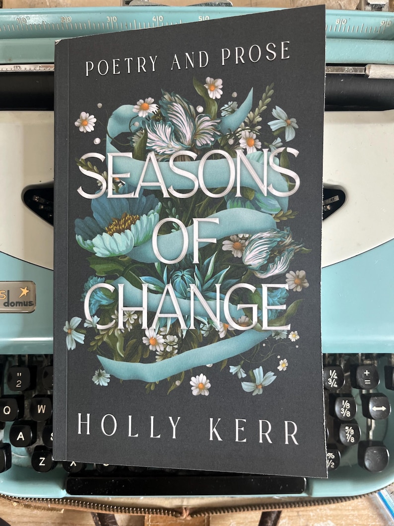 Puede incluir: Un libro titulado "Seasons of Change" de Holly Kerr, con las palabras "Poetry and Prose" en la parte superior. La portada presenta ilustraciones florales en tonos de azul, blanco y verde. El libro descansa sobre una m&aacute;quina de escribir azul vintage.