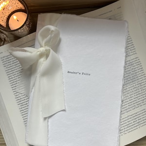 May include: A white paper folio with deckled edges, tied with a cream-colored silk ribbon. The text "Reader's Folio" is printed in black ink on the front cover. The folio rests on top of an open book with visible printed text and pages.
