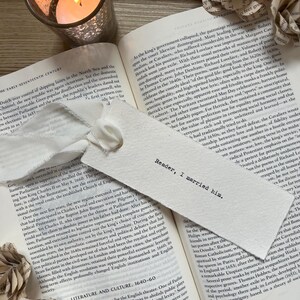 Könnte beinhalten: Ein rechteckiges Lesezeichen aus weißem Papier mit weißem Band und dem schwarzen Aufdruck "Reader, I married him.". Es liegt auf einem aufgeschlagenen Buch, begleitet von einer brennenden Kerze und Papierblumen.