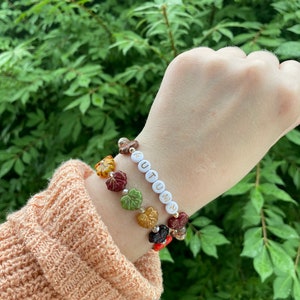 Op de afbeelding: Een armband met witte kralen die "AUTUMN" spellen en drie bedels gemaakt van glazen bladeren in tinten rood, oranje, geel en groen.