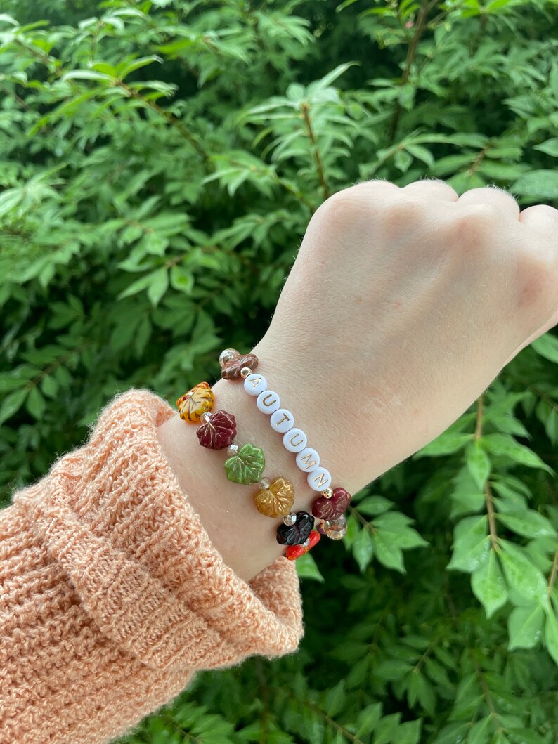 Op de afbeelding: Een armband met witte kralen die "AUTUMN" spellen en drie bedels gemaakt van glazen bladeren in tinten rood, oranje, geel en groen.