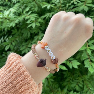 Op de afbeelding: Twee armbanden met herfstthema-charms. De ene armband heeft bruine en witte kralen met een bruine eikel-charm. De andere armband heeft witte kralen met het woord "FALL" gespeld in witte letters.