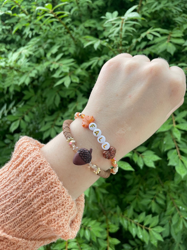 Op de afbeelding: Twee armbanden met herfstthema-charms. De ene armband heeft bruine en witte kralen met een bruine eikel-charm. De andere armband heeft witte kralen met het woord "FALL" gespeld in witte letters.