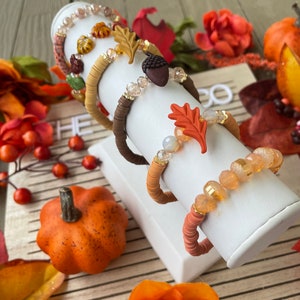 Op de afbeelding: Vijf herfst-thema armbanden met herfstkleuren en bedels, waaronder bladeren, eikels en kralen. De armbanden worden getoond op een witte standaard tegen een houten achtergrond met herfstkleurige bloemen.