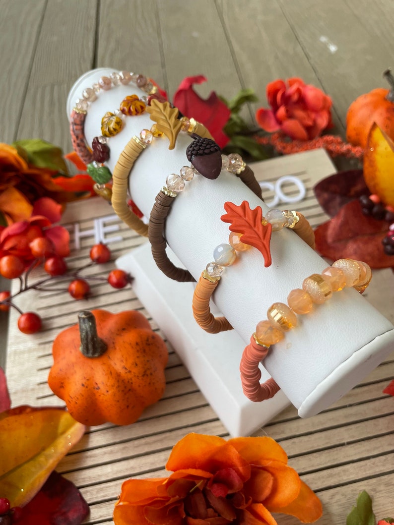Op de afbeelding: Vijf herfst-thema armbanden met herfstkleuren en bedels, waaronder bladeren, eikels en kralen. De armbanden worden getoond op een witte standaard tegen een houten achtergrond met herfstkleurige bloemen.