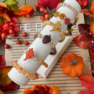 Op de afbeelding: Vijf herfst-thema armbanden met kristallen kralen en bedels, waaronder een eikel, bladeren en een pompoen. De armbanden worden getoond op een witte standaard tegen een houten achtergrond met herfstbladeren.