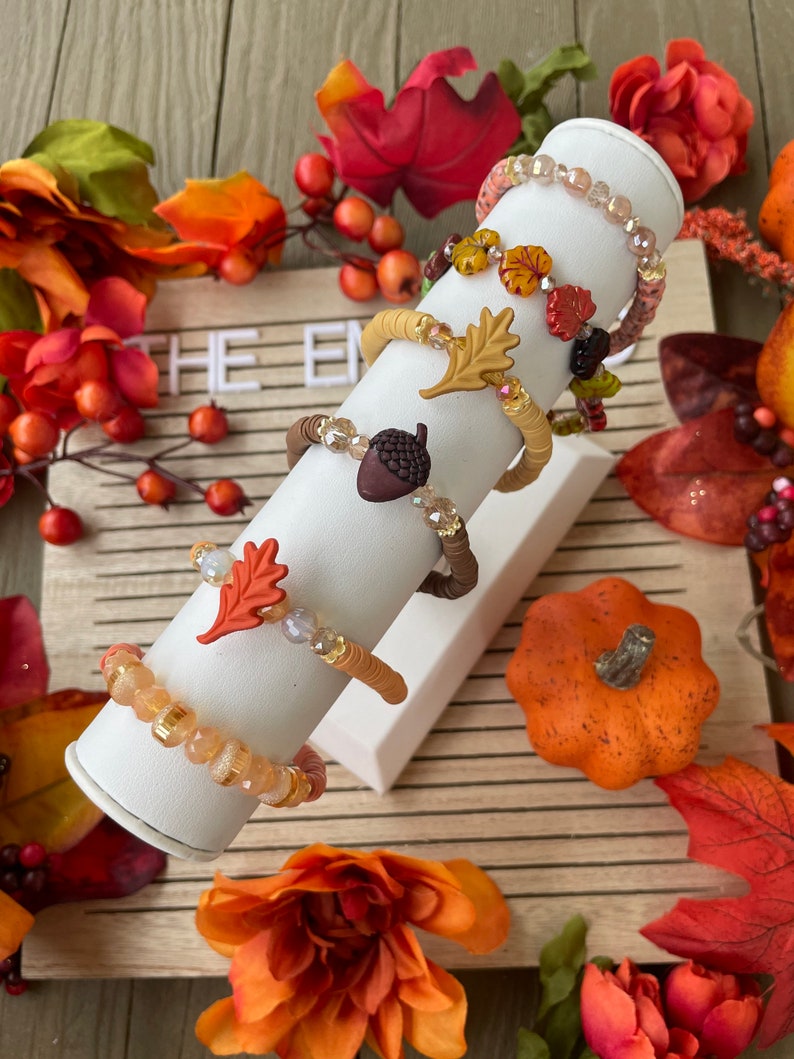 Op de afbeelding: Vijf herfst-thema armbanden met kristallen kralen en bedels, waaronder een eikel, bladeren en een pompoen. De armbanden worden getoond op een witte standaard tegen een houten achtergrond met herfstbladeren.