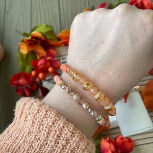 Op de afbeelding: Twee armbanden gestapeld op een pols. De bovenste armband is gemaakt van oranje en gouden kralen. De onderste armband is gemaakt van heldere en bruine kralen.