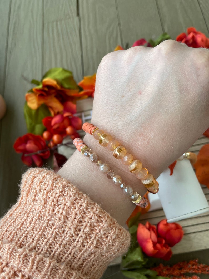 Op de afbeelding: Twee armbanden gestapeld op een pols. De bovenste armband is gemaakt van oranje en gouden kralen. De onderste armband is gemaakt van heldere en bruine kralen.