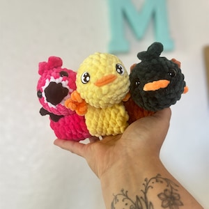 May include: Three crocheted stuffed toys in the shape of birds. The birds are pink, yellow, and black. The pink bird has a white beak and black eyes. The yellow bird has an orange beak and black eyes. The black bird has an orange beak and black eyes.