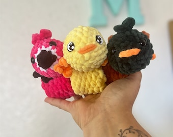 May include: Three crocheted stuffed toys in the shape of birds. The birds are pink, yellow, and black. The pink bird has a white beak and black eyes. The yellow bird has an orange beak and black eyes. The black bird has an orange beak and black eyes.