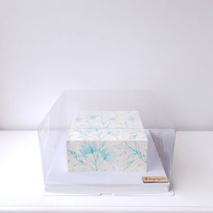 May include: A rectangular cake with a floral design in light blue and white, presented in a clear plastic box. The cake has a delicate pattern of flowers and leaves. The box is on a white surface, with a small wooden sign that reads "All Things Cupcakes".