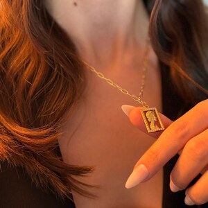 May include: Gold-toned necklace featuring a rectangular pendant with a profile silhouette. The pendant is framed with small, sparkling accents and hangs from a delicate chain. The jewelry is displayed against a dark background.