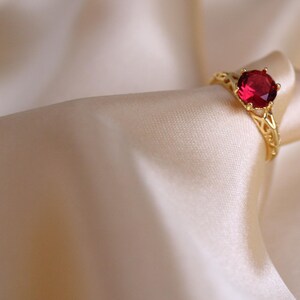 May include: A gold ring featuring a large, round, red gemstone. The ring has an intricate, openwork pattern on the band. It is set against a backdrop of soft, cream-coloured fabric.