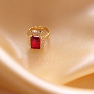 May include: A gold-toned ring with a large, rectangular, red gemstone at the center. The gemstone is surrounded by smaller, clear stones. The ring sits on a gold-colored satin fabric.