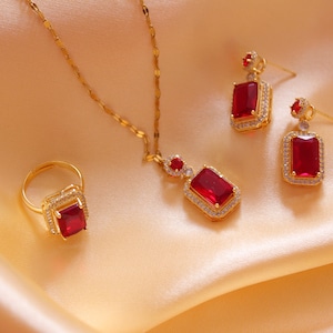 May include: A gold-toned jewelry set featuring a ring, necklace, and earrings. Each piece has a rectangular, deep red gemstone surrounded by small, clear stones. The jewelry rests on a peach-colored, satin-like fabric.