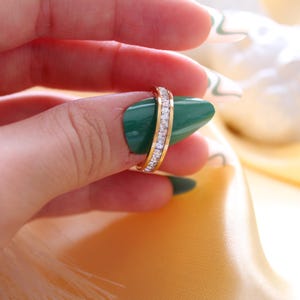 May include: A gold ring with a band of clear, sparkling stones. The ring is held up against a background of yellow fabric and a hand with green nail polish.