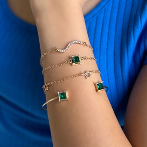 Op de afbeelding: Een verzameling goudkleurige armbanden siert een arm. De ontwerpen omvatten een golvende armband met kleine, heldere stenen, armbanden met vierkante smaragdgroene edelstenen en een met stervormige details. De arm is tegen een blauwe achtergrond.