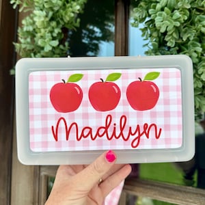 May include: A rectangular, semi-transparent plastic pencil case featuring a label with a light pink and white gingham pattern. The label displays three red apples with green leaves and the name Madilyn written in a red cursive script font.