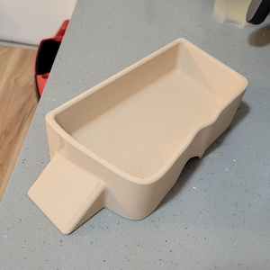 May include: A beige rectangular tray with a handle-like extension on one side. The tray has rounded corners and a smooth surface. The item is likely 3D-printed and designed for holding small items.
