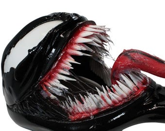 May include: A black, glossy creature head with a large, white, almond-shaped eye and an open mouth filled with sharp, pointed white teeth. A long, red, textured tongue extends from the mouth, which features red, fleshy gums and thin, stringy saliva.