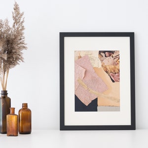 May include: A framed abstract collage with a black frame. The artwork features torn paper pieces in shades of pink, beige, and black. Dried ornamental grass in a brown glass bottle is on the left. Several amber glass bottles are in front.