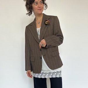 May include: A brown blazer with a subtle striped pattern, paired with black wide-leg pants. The blazer has a decorative pin on the lapel. The outfit includes a white top with lace trim.