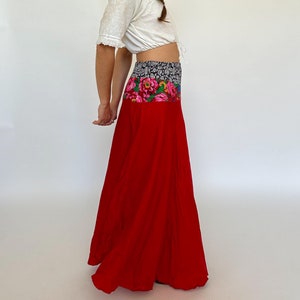 May include: A woman wearing a white crop top and a long red skirt with a floral print waistband. The skirt is flowing and has a relaxed fit.