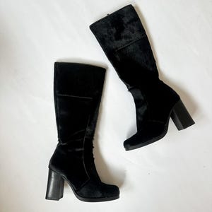 Peut inclure: Une paire de bottes noires montantes avec un aspect texturé et poilu. Les bottes ont un bout carré et un talon bloc épais. Les bottes sont posées sur un fond blanc.