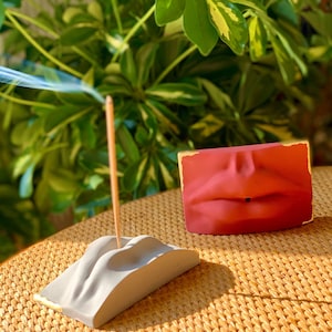 May include: A white ceramic incense holder shaped like a pair of lips with a gold trim. A wooden incense stick is placed in the holder. A red ceramic incense holder shaped like a pair of lips is in the background.