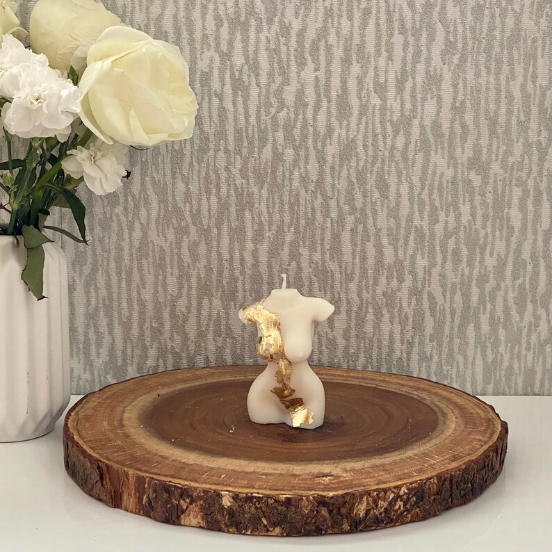 Puede incluir: Vela blanca con forma de figura humana con detalles dorados sobre una rodaja de madera.