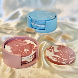 Op de afbeelding: Set van vier roze en witte marmeren onderzetters met een roze houder en een set van vier blauwe en witte marmeren onderzetters met een blauwe houder.