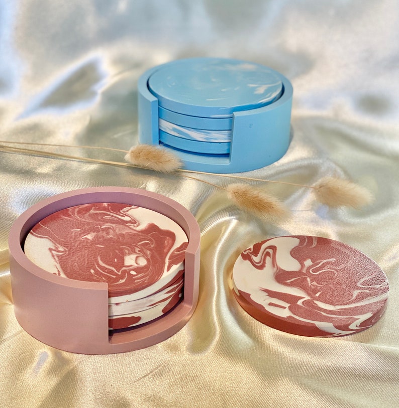 Op de afbeelding: Set van vier roze en witte marmeren onderzetters met een roze houder en een set van vier blauwe en witte marmeren onderzetters met een blauwe houder.