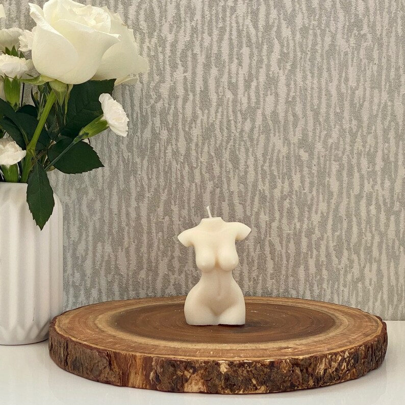 Puede incluir: Una vela blanca con forma de torso humano, sentada sobre una rodaja de madera. La vela est&aacute; sobre un fondo blanco.