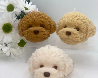 Puede incluir: Tres velas con forma de perro, una marrón, una beige y una blanca. Las velas tienen todas la forma de la cabeza de un caniche.